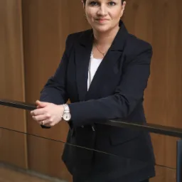 Portret kobiety w granatowym garniturze, opierającej się o czarną barierkę, z zegarkiem na nadgarstku i delikatną biżuterią, na tle drewnianej ściany.