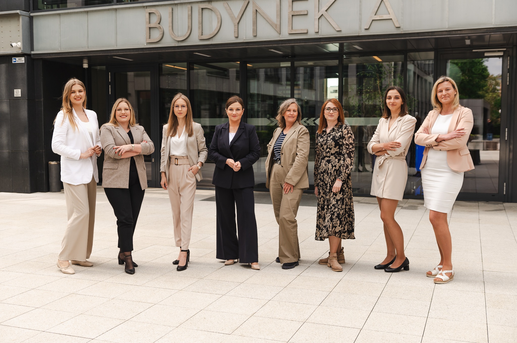 Grupa siedmiu kobiet w różnym wieku, ubranych w stroje biznesowe, stojących przed nowoczesnym budynkiem biurowym z napisem 'BUDYNEK A' nad wejściem.
