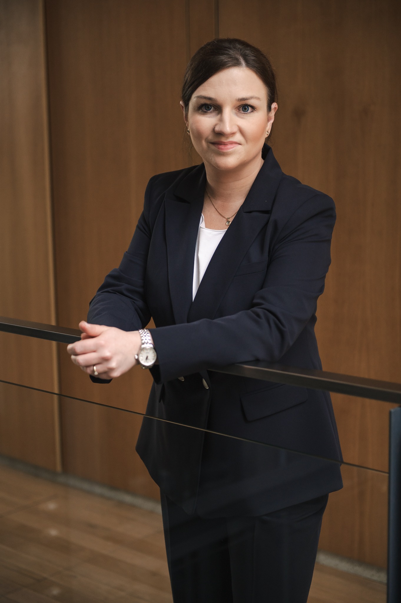 Portret kobiety w granatowym garniturze, opierającej się o czarną barierkę, z zegarkiem na nadgarstku i delikatną biżuterią, na tle drewnianej ściany.