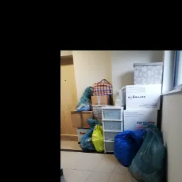 Stos kartonów, toreb i koszyków z rzeczami w korytarzu, przygotowanych do przeniesienia, z widocznymi napisami na kartonach.