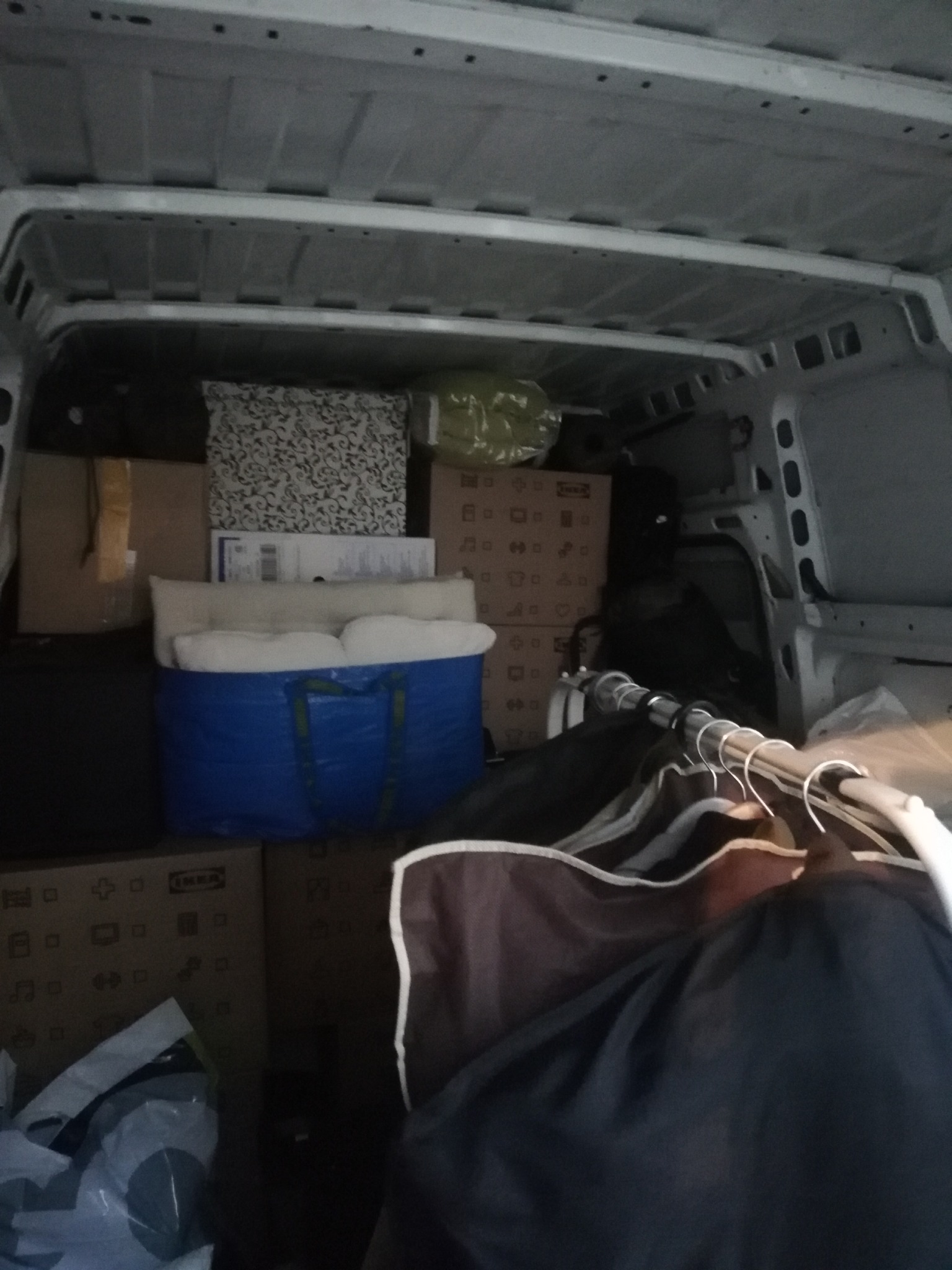 Wnętrze furgonetki załadowane kartonami, torbami IKEA i ubraniami na wieszakach, przygotowane do transportu.