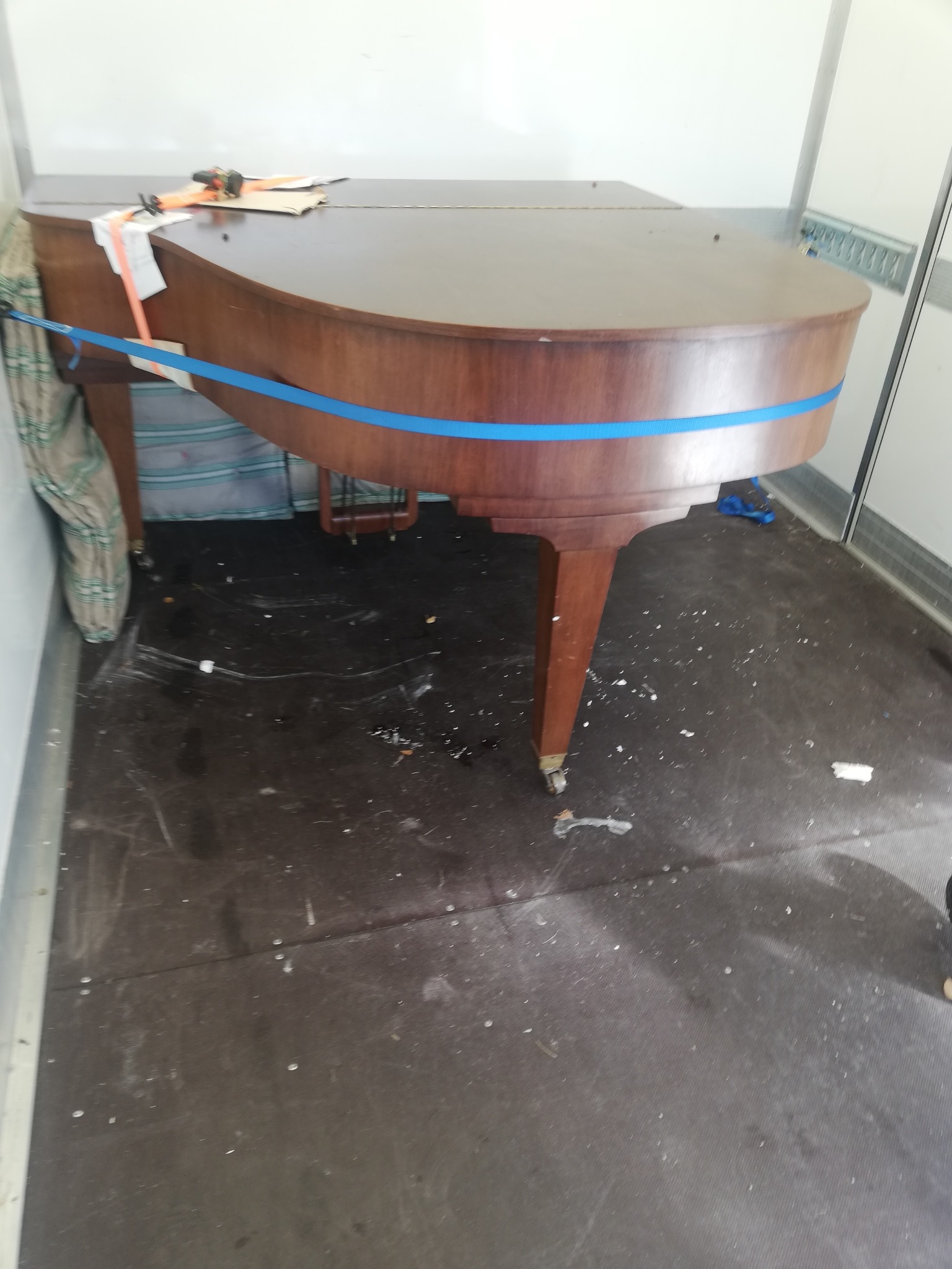 Pianino zabezpieczone pasami transportowymi wewnątrz pojazdu dostawczego, przygotowane do transportu, z widocznymi śladami po przeprowadzce na podłodze.