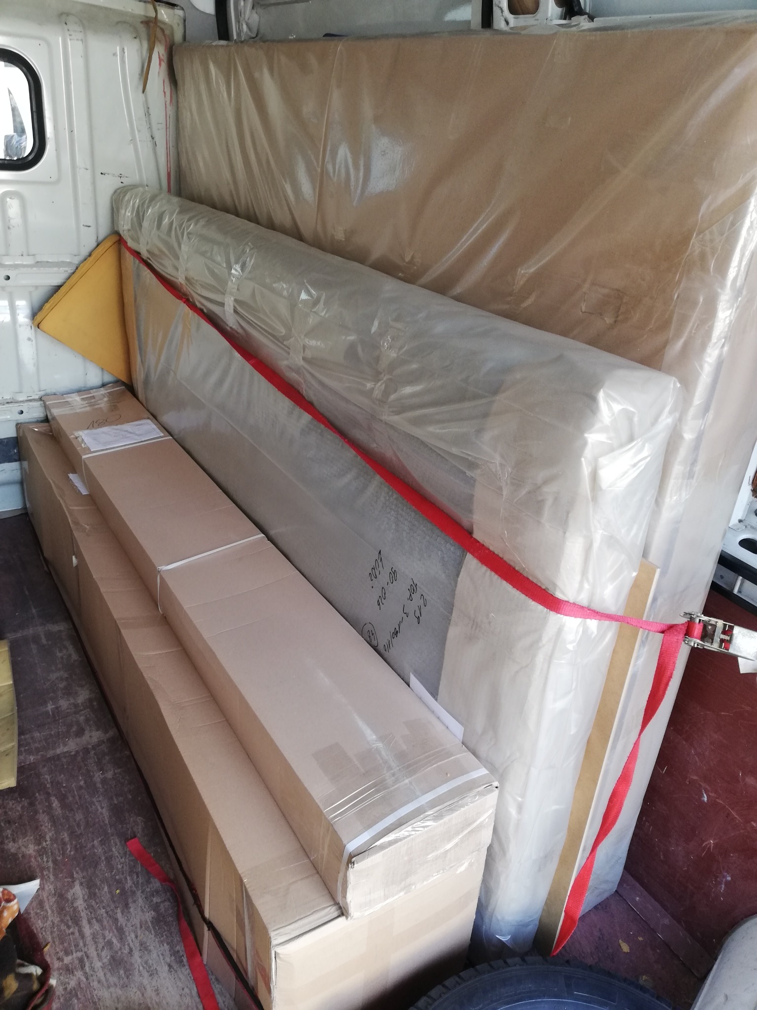 Wnętrze furgonetki załadowane kartonami i meblami owiniętymi folią, zabezpieczone czerwoną taśmą transportową.
