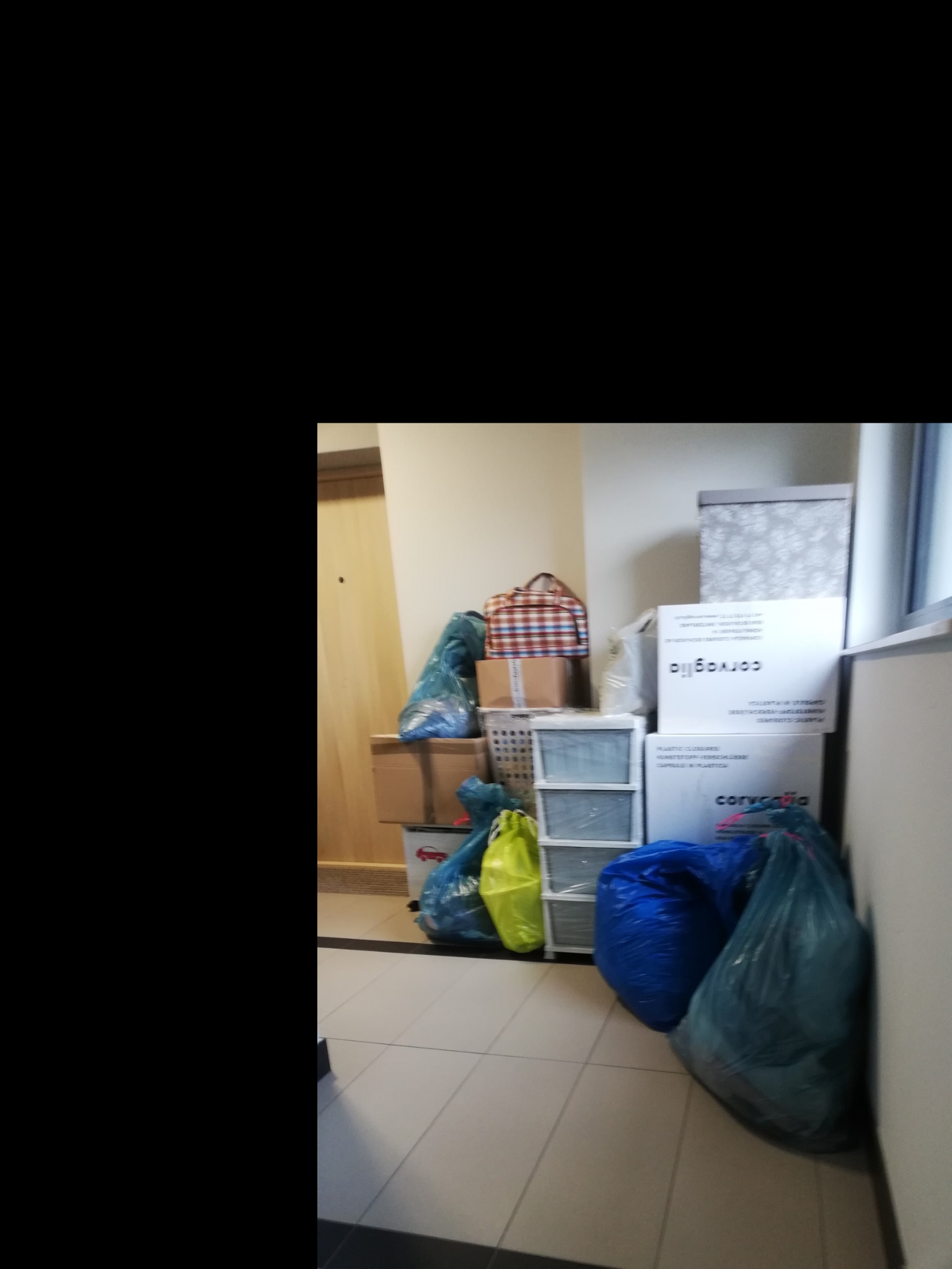 Stos kartonów, toreb i koszyków z rzeczami w korytarzu, przygotowanych do przeniesienia, z widocznymi napisami na kartonach.