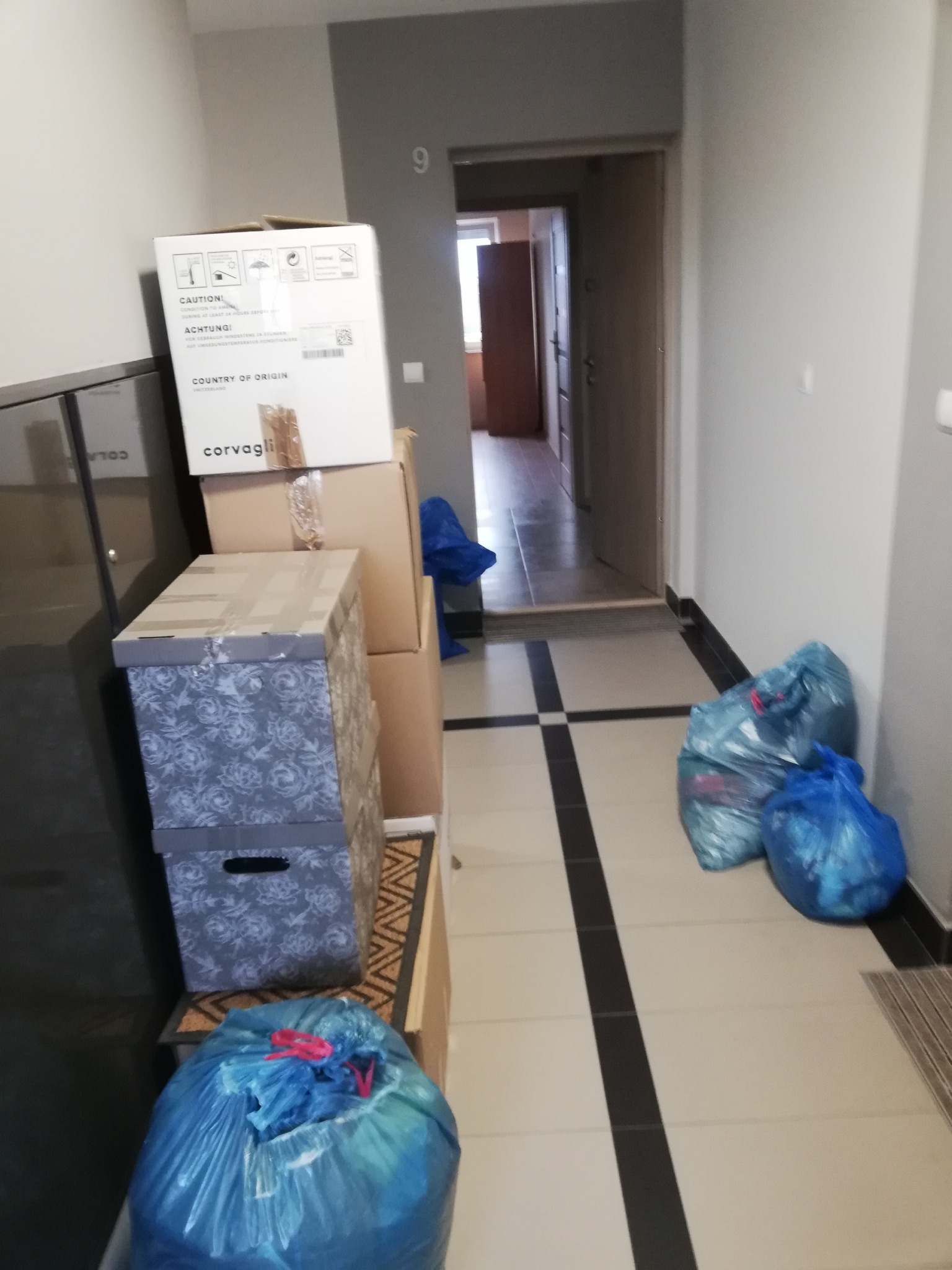 Korytarz mieszkania z przygotowanymi kartonami i workami do spakowania, widoczny numer mieszkania '9' na ścianie.