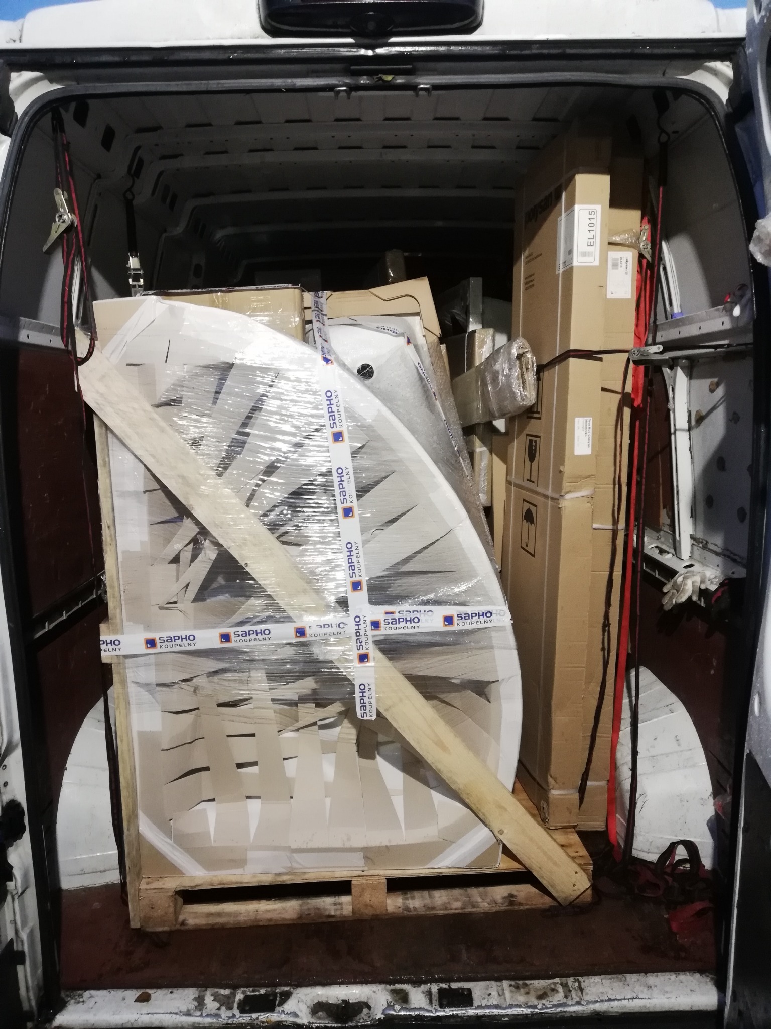 Wnętrze białego busa wypełnione paletą z owalną kabiną prysznicową marki SAPHO, zabezpieczoną folią stretch i drewnianymi belkami, oraz kartonami różnych rozmiarów, widoczne pasy mocujące...
