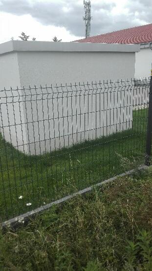 Ciemna, metalowa siatka ogrodzeniowa, widok na trawnik i budynek z jasną elewacją oraz wieżę telekomunikacyjną w tle.