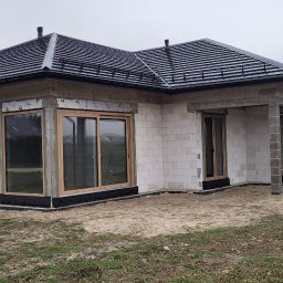 Okno-Pol Okna ,Drzwi, Rolety ,Bramy Pińczów, Busko-Zdrój , Jedrzejów ,Chmielnik - Dom w trakcie budowy z zamontowanymi oknami z drewnianą ramą. Widoczne bloczki betonowe i czarna dachówka. Na zewnątrz piasek i trawa.