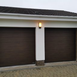 Okno-Pol Okna ,Drzwi, Rolety ,Bramy Pińczów, Busko-Zdrój , Jedrzejów ,Chmielnik - Zamknięte, brązowe, segmentowe drzwi garażowe z poziomymi panelami, oświetlone lampą zewnętrzną. Widoczny fragment dachu i kostka brukowa.