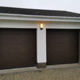 Zamknięte, brązowe, segmentowe drzwi garażowe z poziomymi panelami, oświetlone lampą zewnętrzną. Widoczny fragment dachu i kostka brukowa.