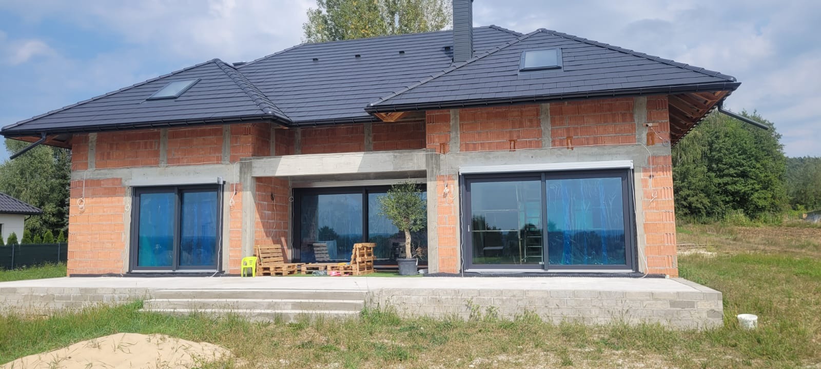 Surowy stan domu jednorodzinnego z grafitową dachówką, widoczne okna przesuwne z folią ochronną i niezabudowana elewacja z cegły. W tle zieleń drzew.
