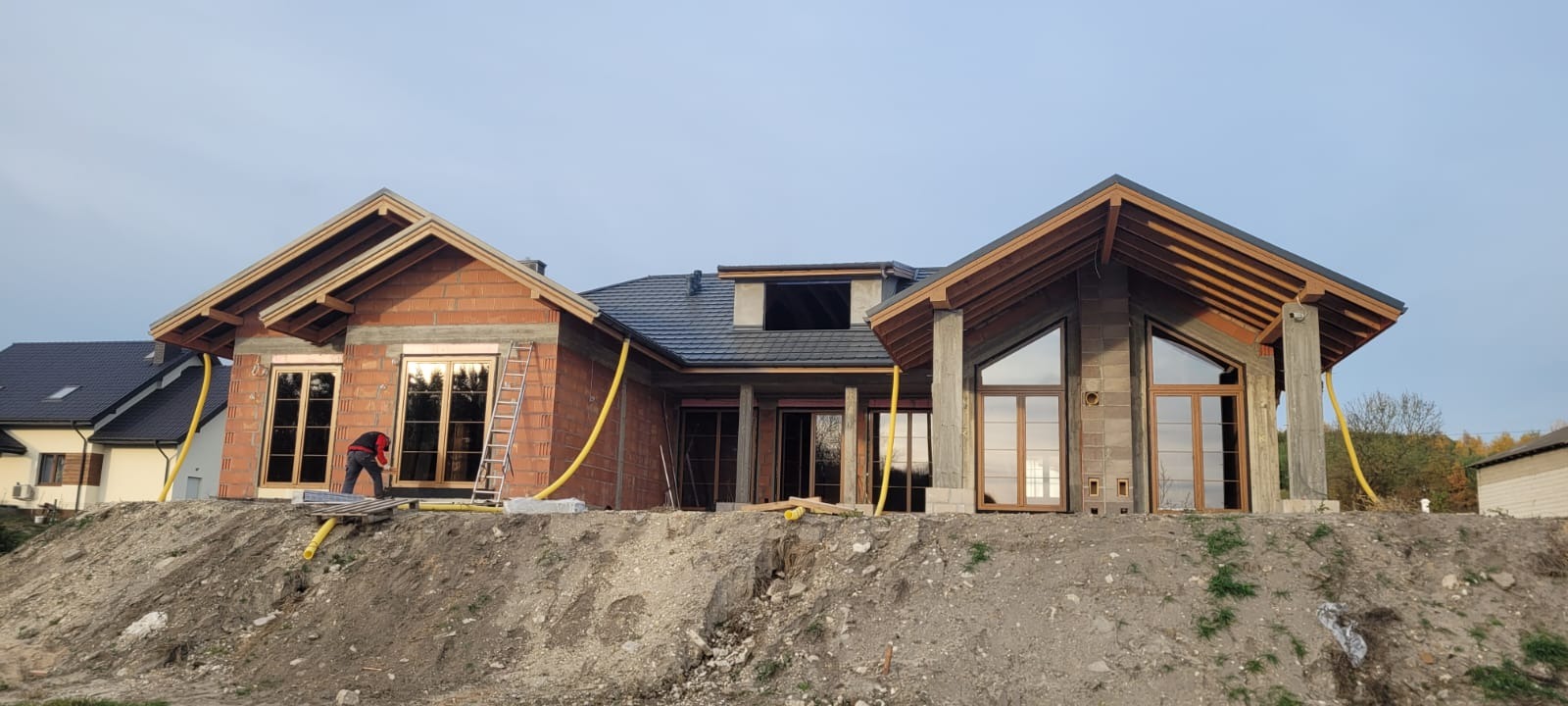 Budowa domu jednorodzinnego w stanie surowym otwartym. Widoczne okna z brązowymi ramami, cegły, pustaki i żółty wąż. Na pierwszym planie usypana ziemia.