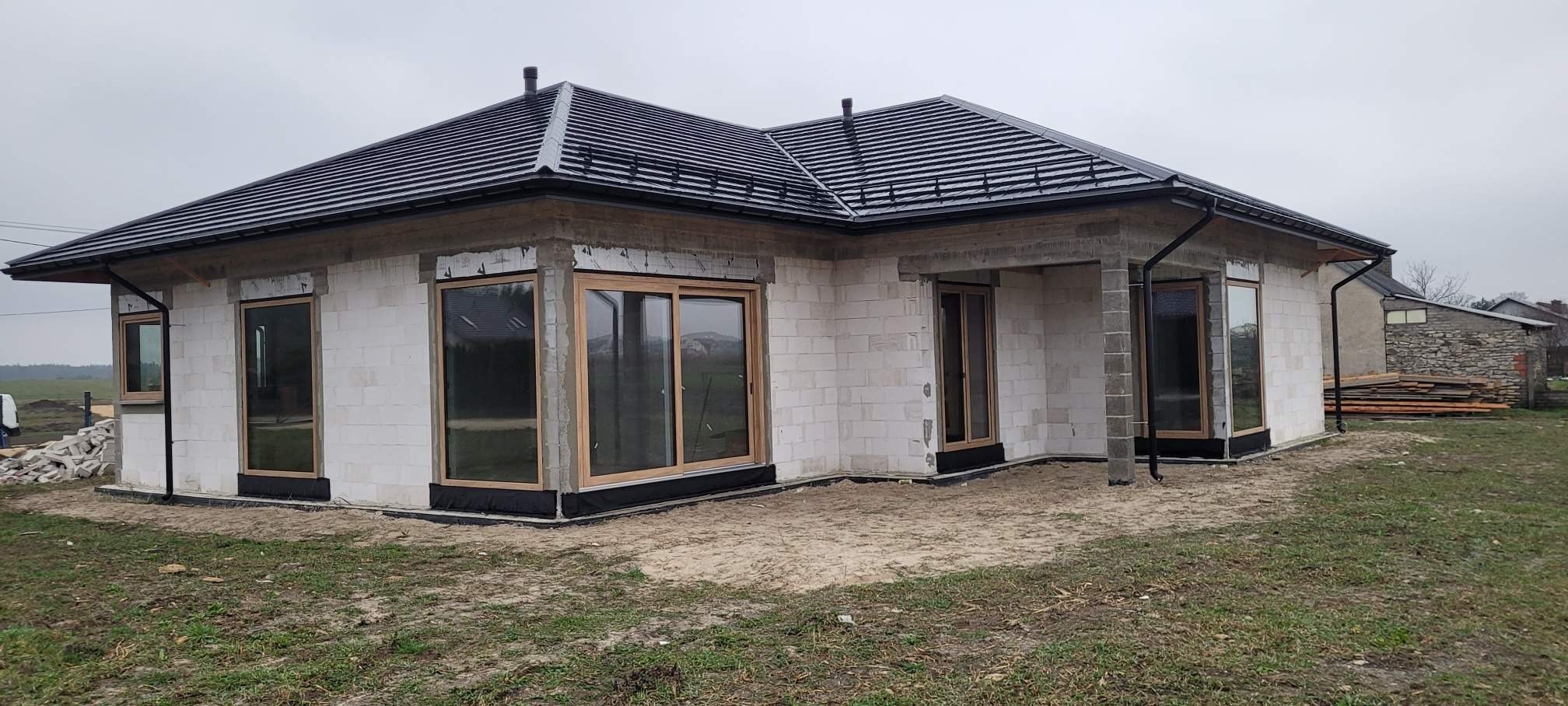 Dom w trakcie budowy z zamontowanymi oknami z drewnianą ramą. Widoczne bloczki betonowe i czarna dachówka. Na zewnątrz piasek i trawa.