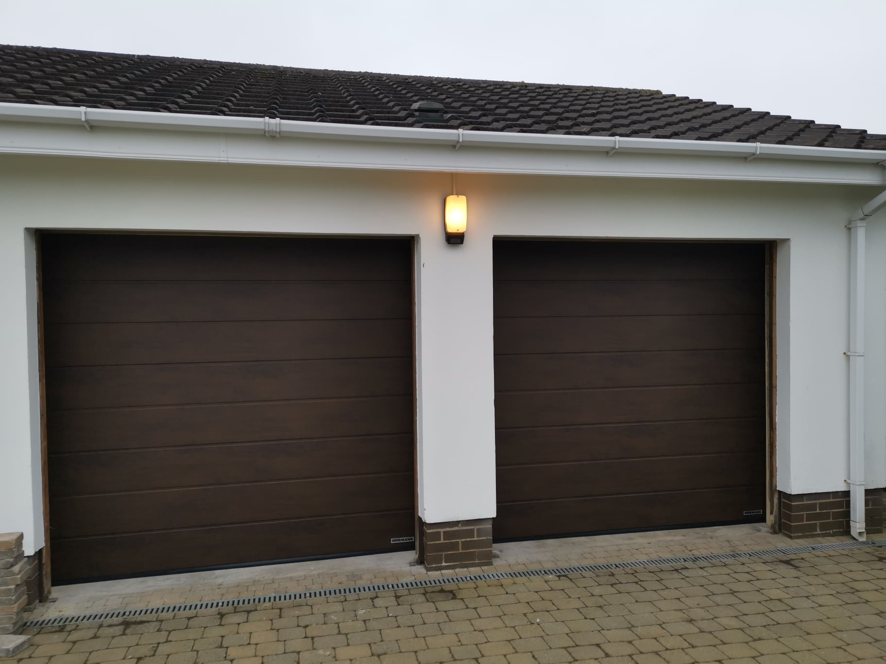 Zamknięte, brązowe, segmentowe drzwi garażowe z poziomymi panelami, oświetlone lampą zewnętrzną. Widoczny fragment dachu i kostka brukowa.