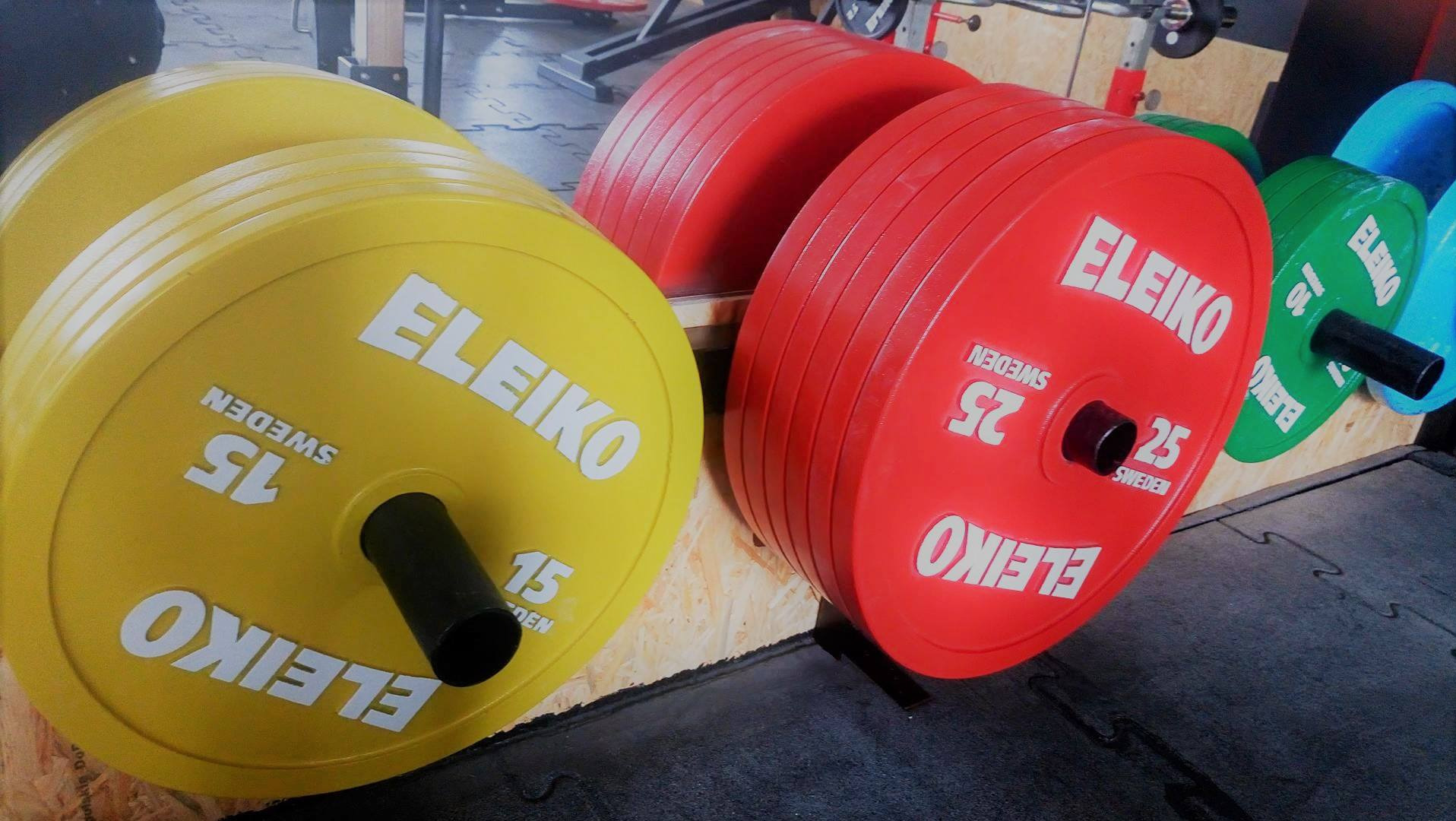 Kolorowe obciążenia do sztangi marki Eleiko: żółte 15 kg, czerwone 25 kg, zielone i niebieskie, ułożone na stojaku w siłowni.