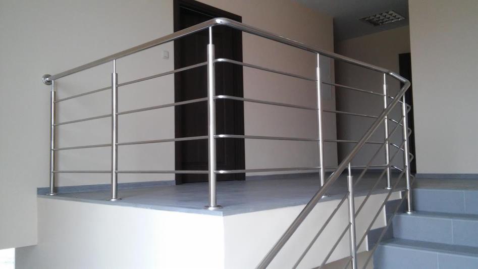 Wewnętrzna klatka schodowa z nowoczesną, minimalistyczną balustradą ze stali nierdzewnej z poziomymi prętami, widok z dołu na schody wyłożone szarą płytką.