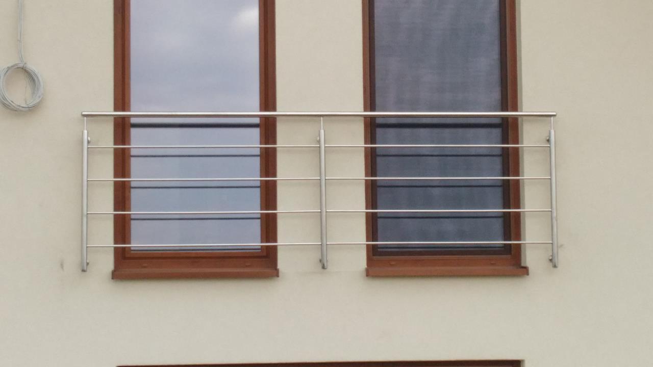 Nierdzewna balustrada okienna z poziomymi prętami, zamontowana na elewacji budynku z brązowymi ramami okiennymi, widoczne zwisające kable po lewej stronie.