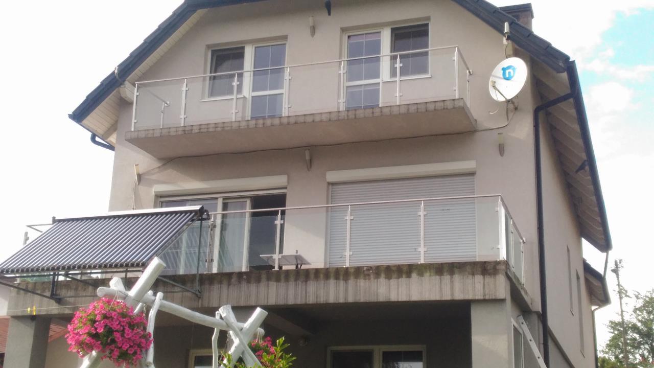 Dwukondygnacyjny dom z balkonami zabezpieczonymi balustradami szklanymi z białymi słupkami, widok z dołu na elewację z anteną satelitarną i kolektorami słonecznymi.