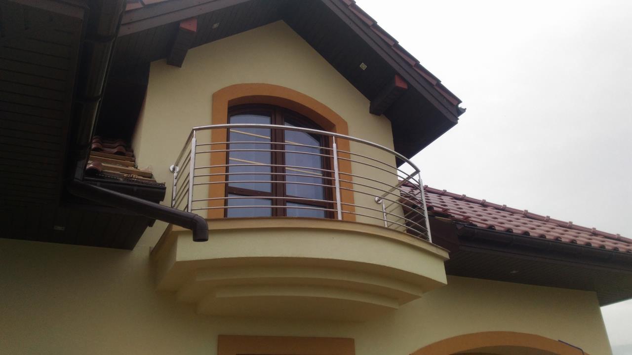 Balkon z zaokrągloną, nierdzewną balustradą z poziomych prętów, na tle okna z brązową ramą w budynku z beżową elewacją i brązowym dachem.