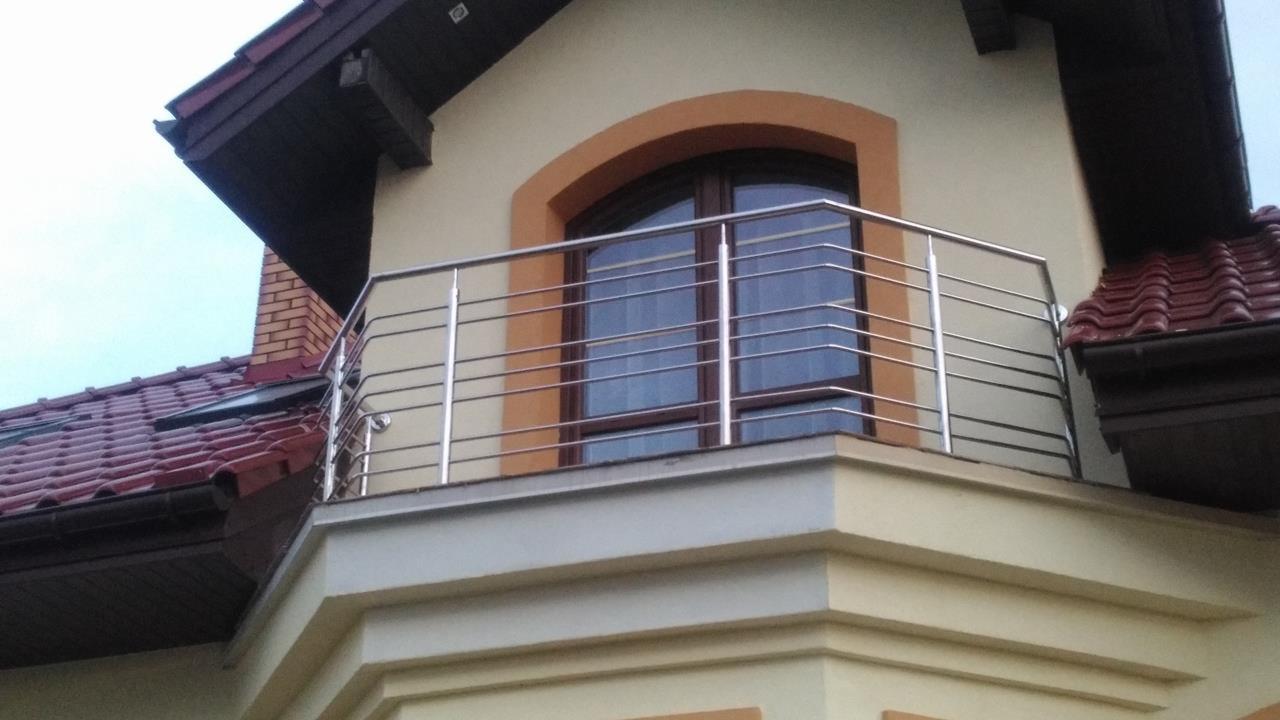 Balkon z nowoczesną, poziomą balustradą ze stali nierdzewnej, widok z dołu na tle okna z łukowatym obramowaniem i dachu z czerwonej dachówki.