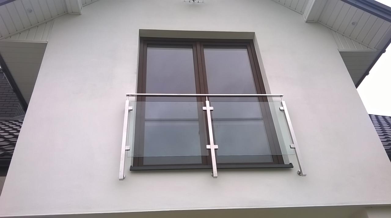 Szklana balustrada balkonowa z nierdzewnymi elementami mocującymi, widoczna z zewnątrz, na tle białej elewacji domu z brązowymi oknami.