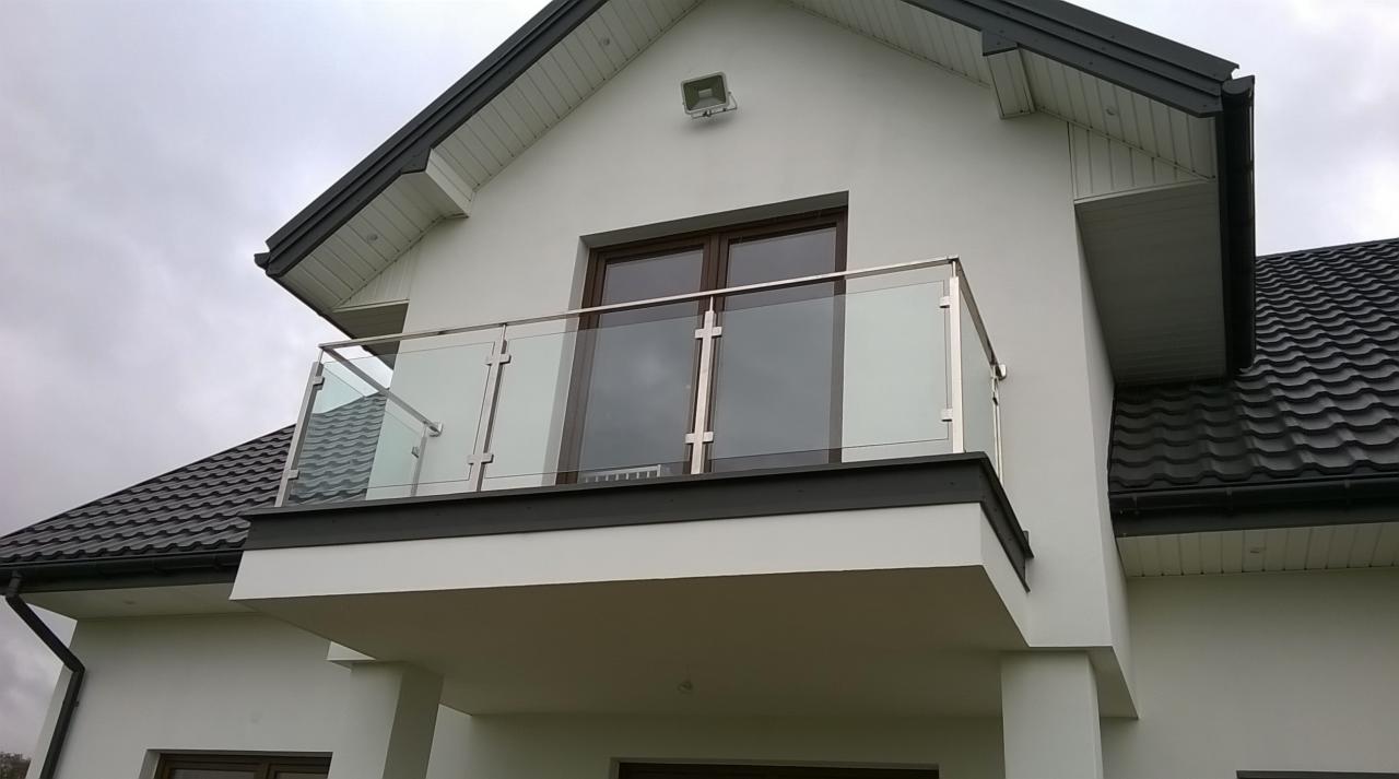 Balkon z nowoczesną szklaną balustradą z elementami ze stali nierdzewnej, widok z dołu na tle białej elewacji domu z ciemną dachówką.