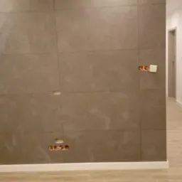 Ściana wyłożona szarymi, matowymi płytkami imitującymi kamień, z widocznymi otworami na gniazdka elektryczne i puszkami instalacyjnymi, w nowym, jasnym wnętrzu z drewnopodobną podłogą i białymi...