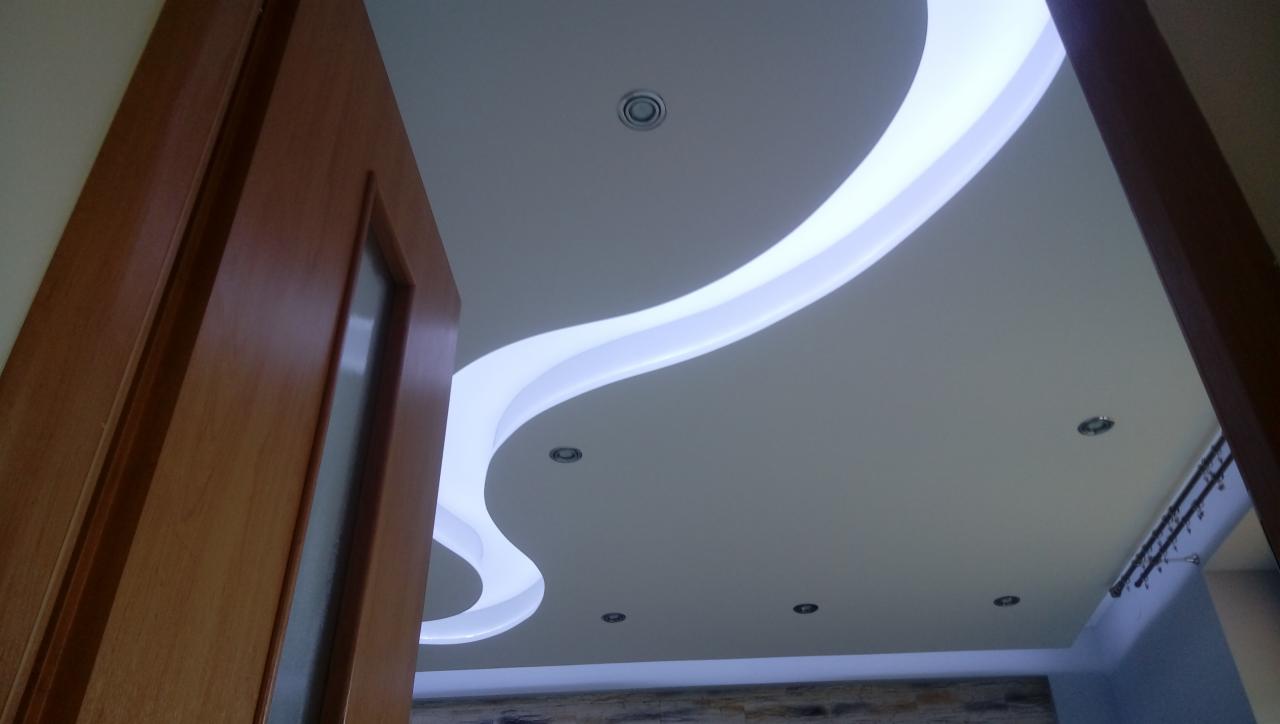 Nowoczesny sufit podwieszany z falistym oświetleniem LED i punktowymi lampami, widoczny fragment drewnianej ościeżnicy drzwi.