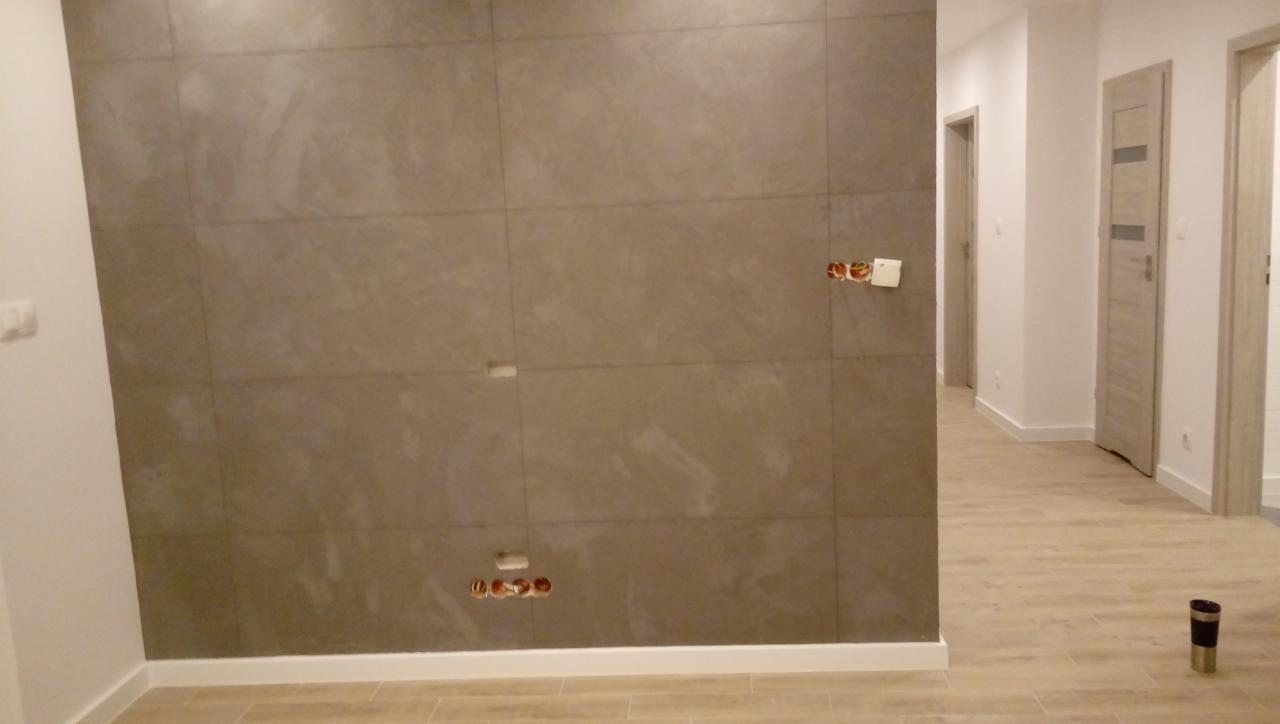 Ściana wyłożona szarymi, matowymi płytkami imitującymi kamień, z widocznymi otworami na gniazdka elektryczne i puszkami instalacyjnymi, w nowym, jasnym wnętrzu z drewnopodobną podłogą i białymi...