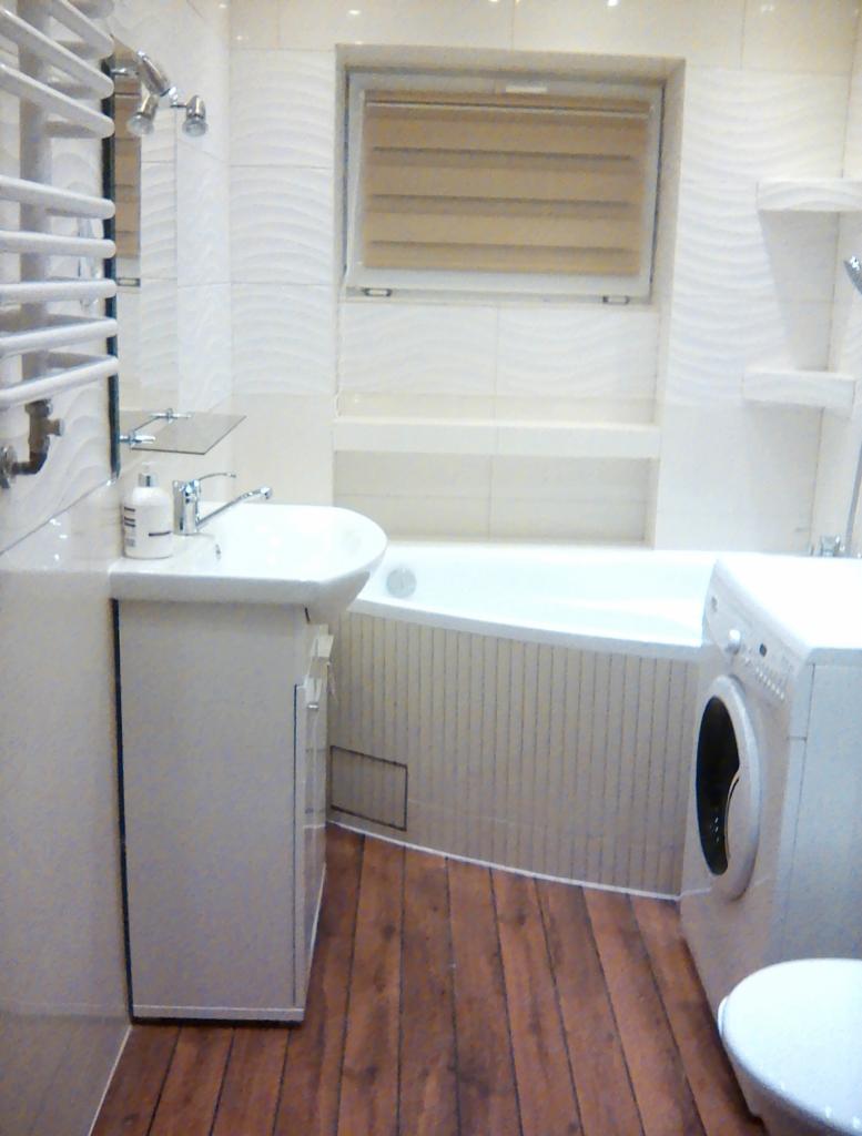 Jasna łazienka z drewnopodobną podłogą, białą armaturą i pralką; widoczny grzejnik drabinkowy, umywalka z szafką, wanna obudowana płytkami oraz okno z żaluzjami.