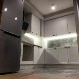 Nowoczesna kuchnia z białymi szafkami, podświetleniem LED pod górnymi szafkami i szarą lodówką Samsung, widok z niskiego kąta.