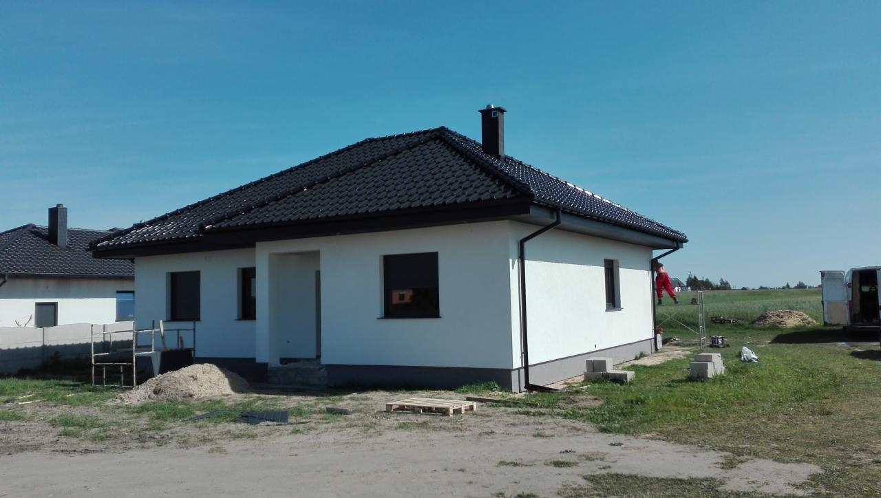 Nowoczesny dom jednorodzinny z ciemną dachówką, widoczny fragment drugiego domu w tle, pracownik w czerwonym kombinezonie przy rynnie, paleta i gruz na trawniku.