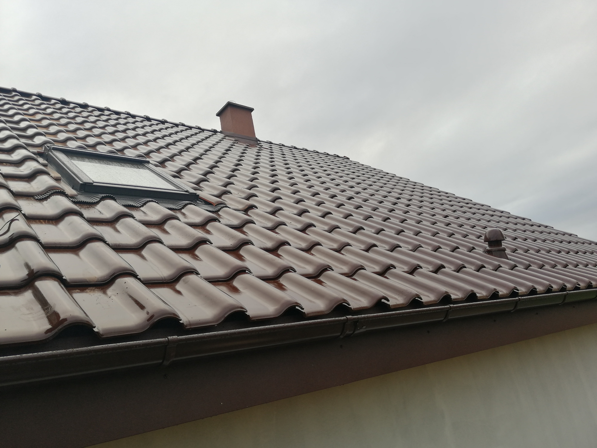 Ciemnobrązowy dach z dachówką ceramiczną, widoczne okno dachowe, komin i rynna w pochmurny dzień.