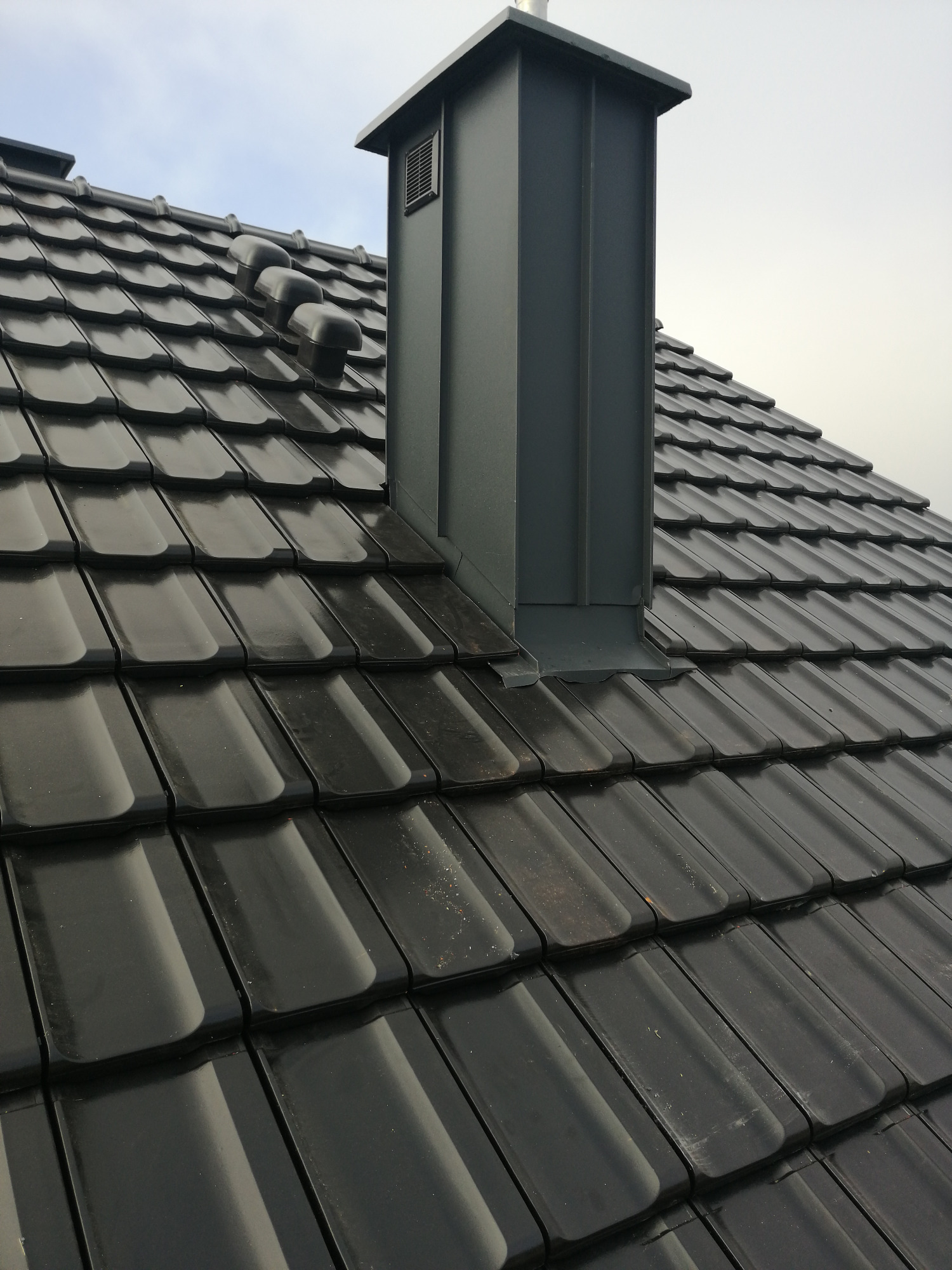 Błyszczące, czarne dachówki ceramiczne z trzema kominkami wentylacyjnymi i nowoczesnym, metalowym kominem w kolorze grafitowym na tle pochmurnego nieba.