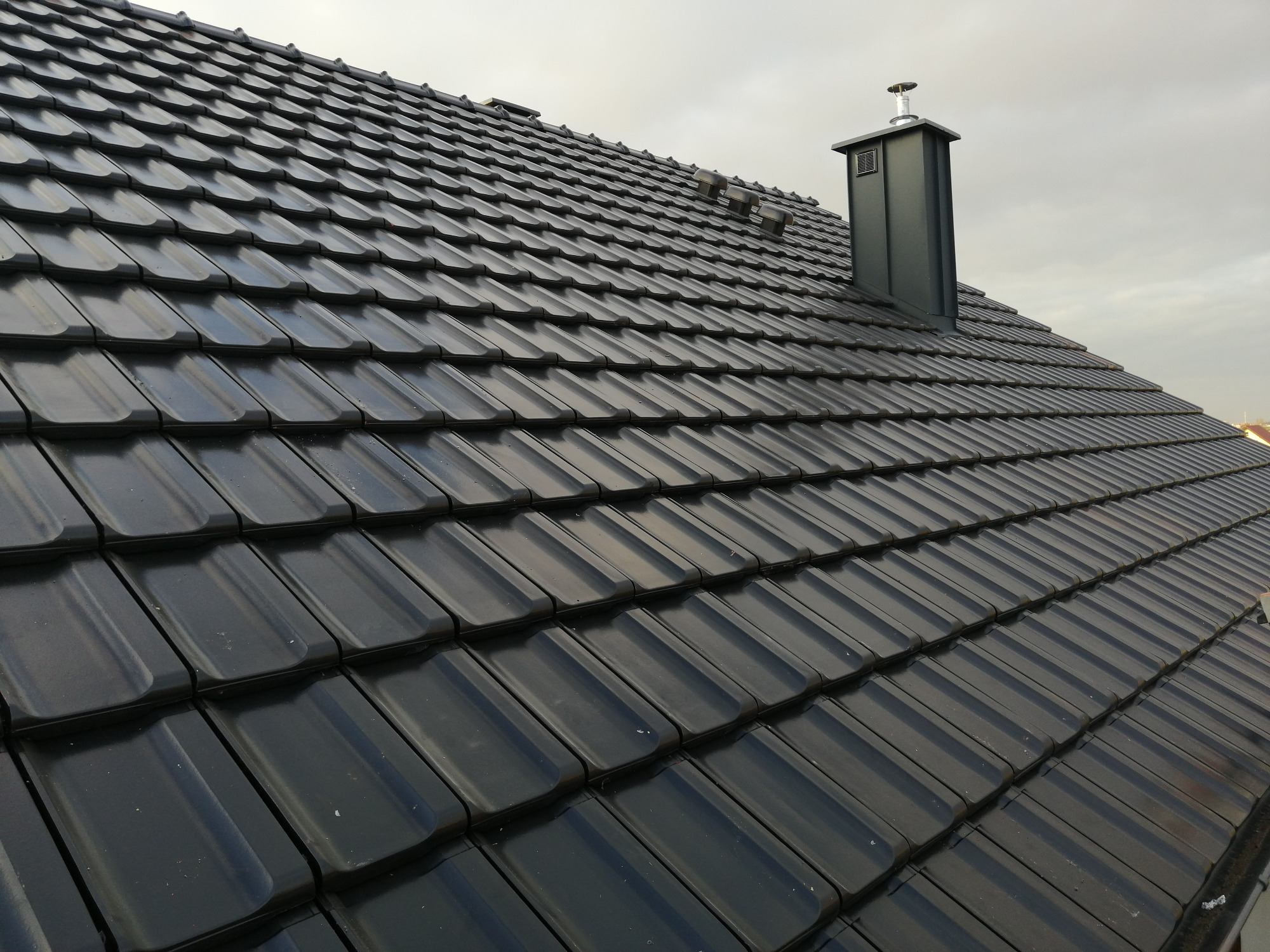 Czarna, błyszcząca dachówka ceramiczna ułożona na nowym dachu, z widocznym kominem wentylacyjnym i elementami wentylacji kalenicy, sfotografowana pod kątem z dołu.