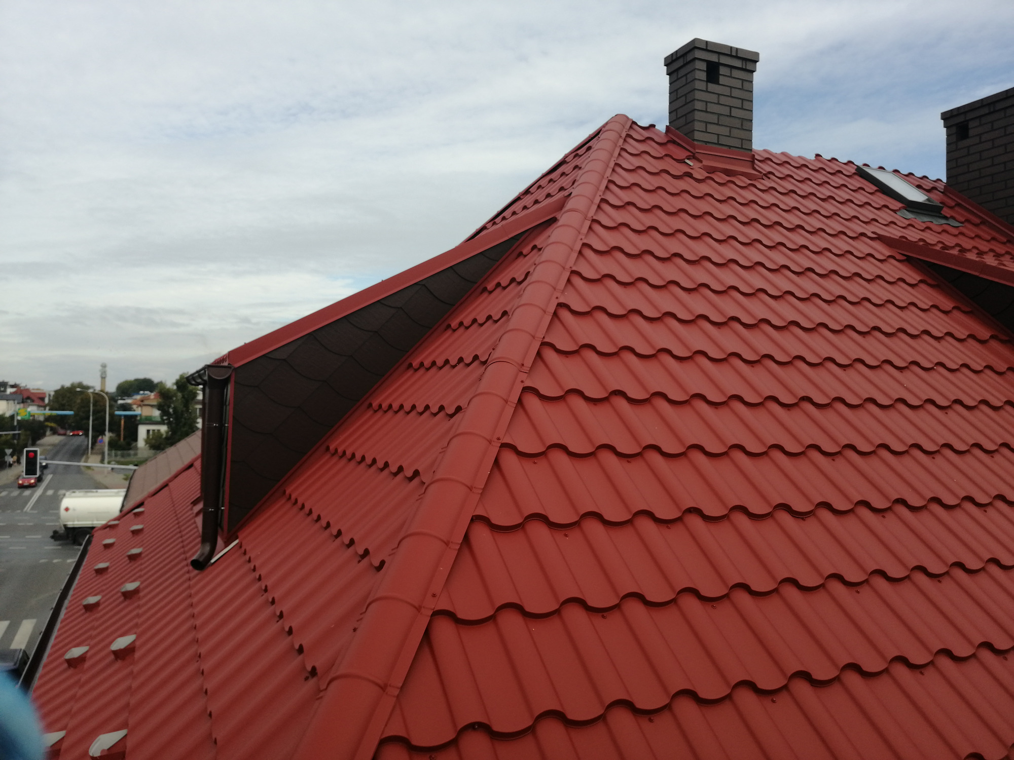 Czerwony dach z blachy trapezowej z dwoma kominami z cegły i oknem dachowym, widok z góry na tle pochmurnego nieba.