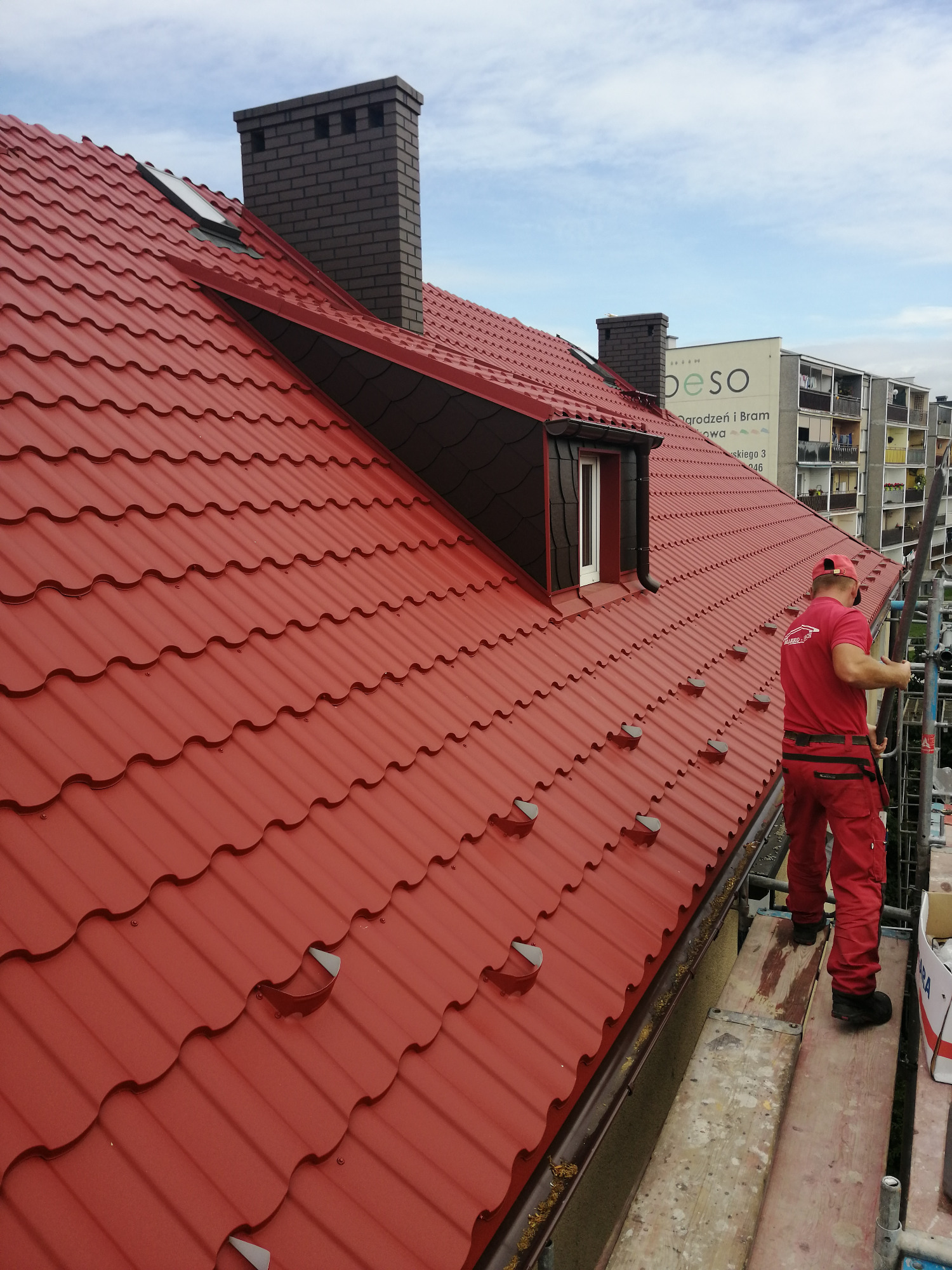 Czerwony dach z blachodachówki trapezowej, komin z cegły klinkierowej, okno dachowe i dekarz w czerwonym kombinezonie na rusztowaniu podczas prac.