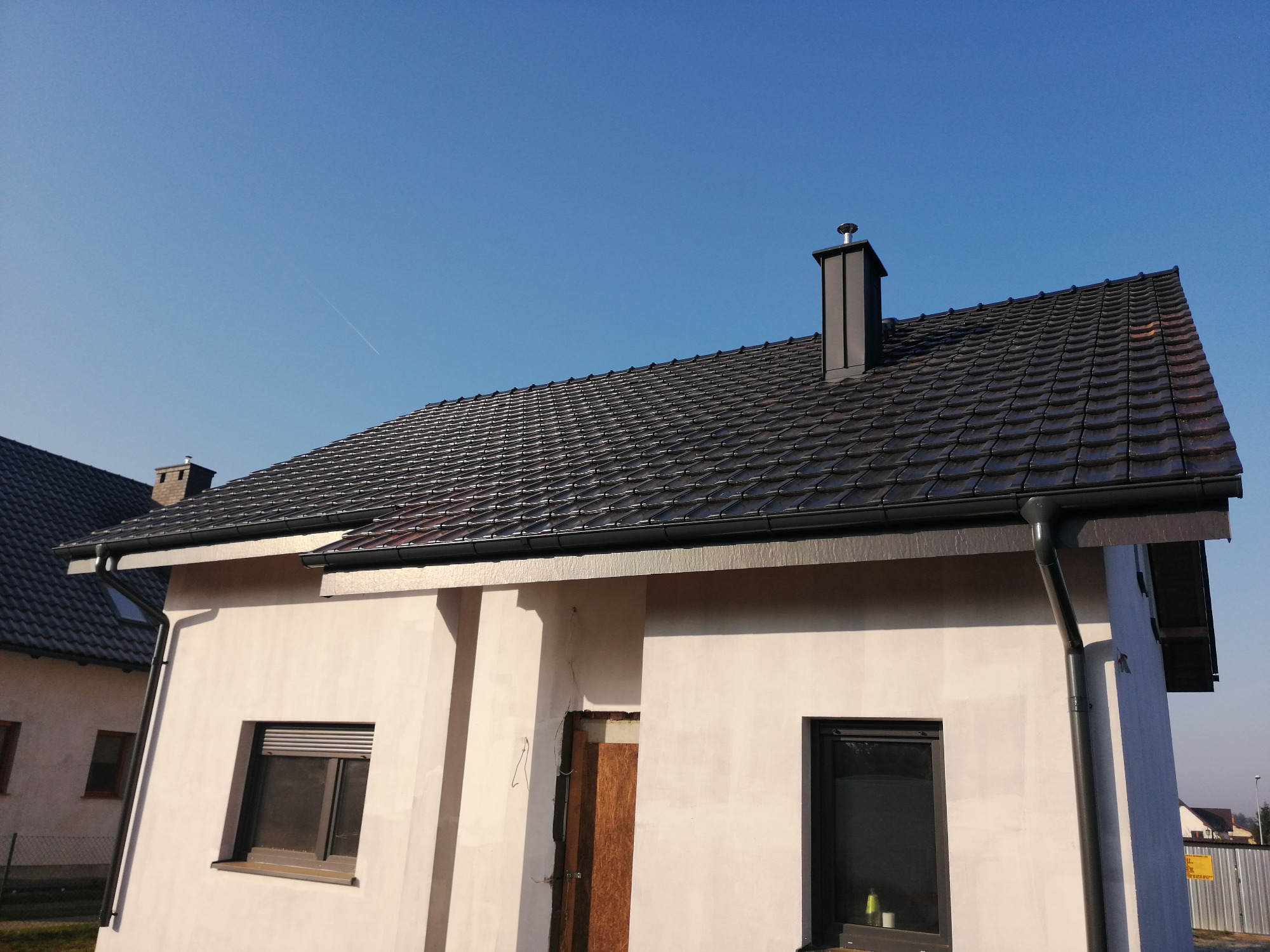 Nowy dach z ciemnej dachówki ceramicznej na domu jednorodzinnym w trakcie wykończenia, widoczne czarne rynny i komin na tle błękitnego nieba.
