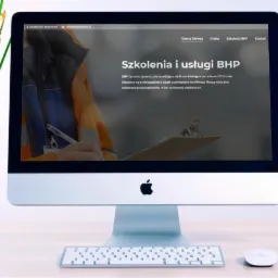 Ekran komputera iMac wyświetlający stronę internetową z ofertą szkoleń i usług BHP, obok doniczka z kwitnącymi żonkilami.