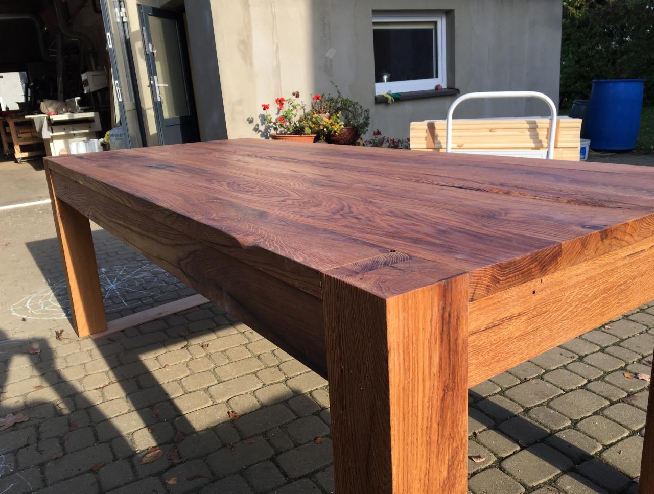 Solidny stół z ciemnego drewna na grubych nogach, widoczne słoje i naturalne wykończenie, ustawiony na brukowanym podwórku w słoneczny dzień.