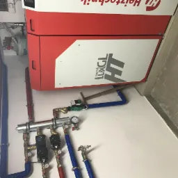 Czerwono-biały piec z podłączonymi rurami miedzianymi i niebieskimi, manometry i zawory widoczne na białej ścianie w kotłowni.