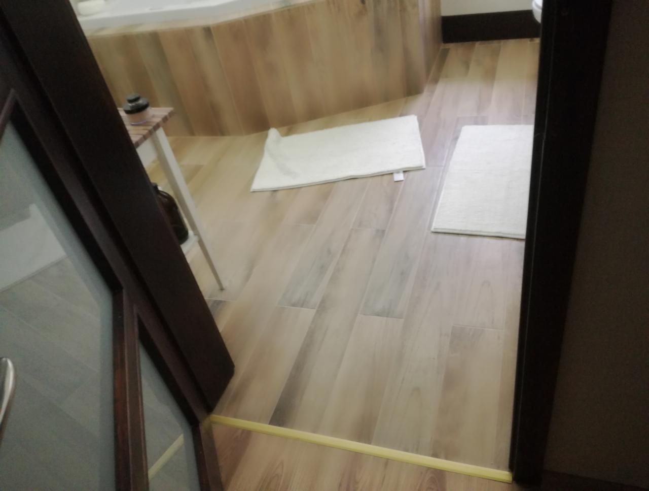 Widok zza uchylonych drzwi na łazienkę z drewnopodobnymi płytkami podłogowymi, dwoma białymi dywanikami, fragmentem wanny obłożonej płytkami i małym stolikiem z kosmetykami.