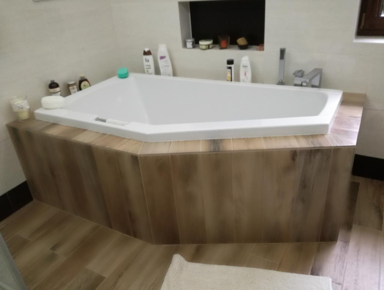 Biała, narożna wanna obudowana płytkami imitującymi drewno, z widoczną armaturą i kosmetykami, na podłodze płytki w podobnym stylu, z fragmentem białego dywanika.