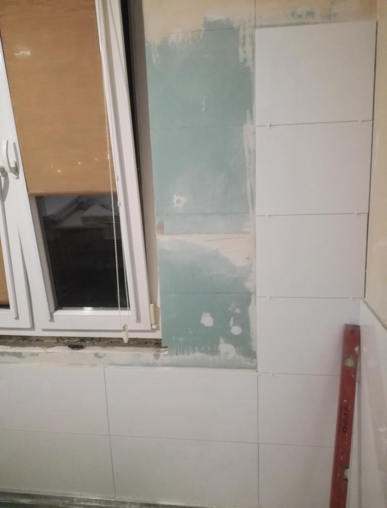 Układanie białych płytek ściennych w łazience przy oknie, widoczny fragment ściany przygotowanej pod płytki i poziomica przy ścianie.
