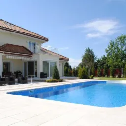 Nowoczesny dom z dużymi oknami i przesuwnymi drzwiami tarasowymi, widok z perspektywy basenu w słoneczny dzień.