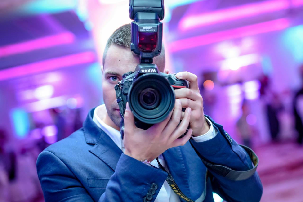 Mężczyzna w niebieskiej marynarce celuje obiektywem aparatu Nikon z lampą błyskową wprost w widza na tle rozmytych świateł sali.