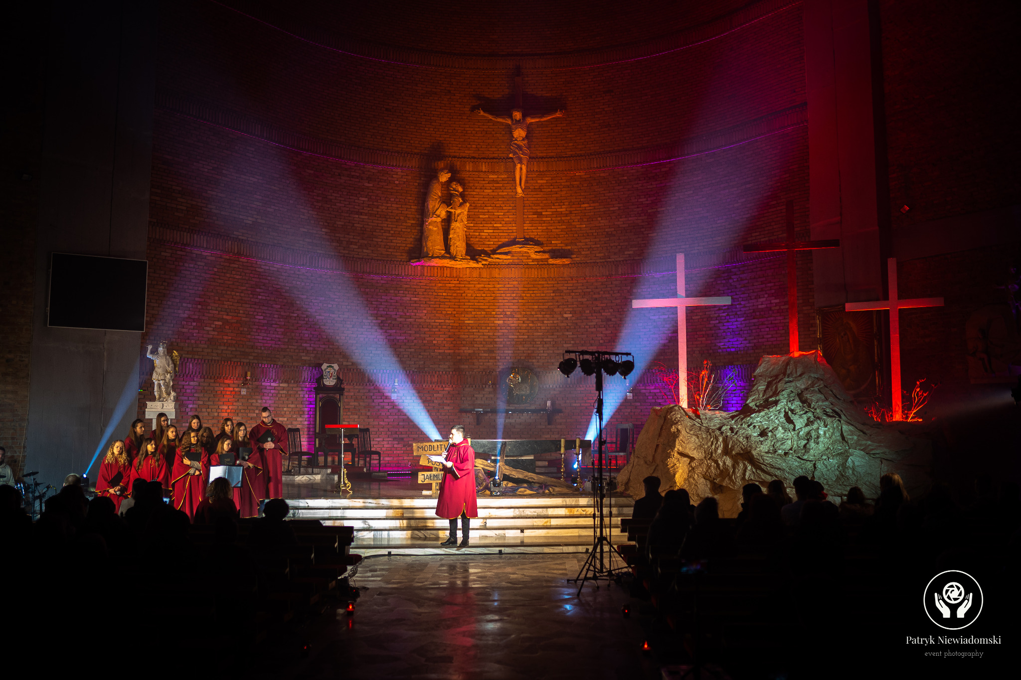 Występ chóru w czerwonych szatach na scenie w kościele, oświetlonej reflektorami i dekoracjami tematycznymi, w tle krzyż i ceglana ściana.