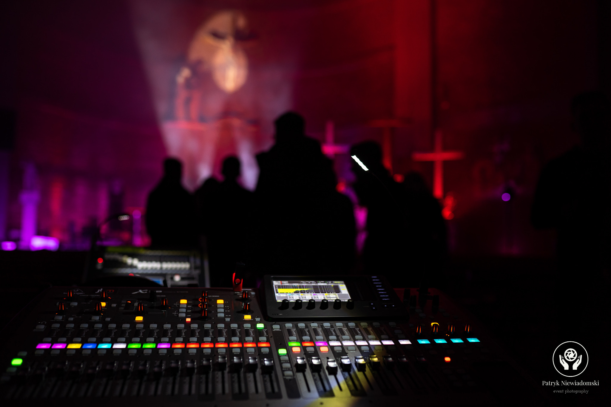 Profesjonalny mikser dźwięku z podświetlanymi przyciskami i suwakami, ustawiony na tle rozmytej sceny z sylwetkami osób i efektami świetlnymi w odcieniach czerwieni i fioletu.