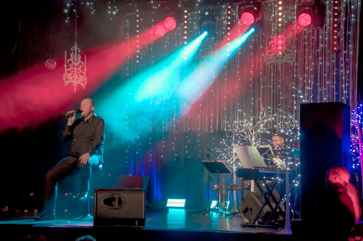 Wokalista śpiewa do mikrofonu na scenie z dekoracjami świetlnymi, za nim muzyk gra na keyboardzie, a w tle czerwono-niebieskie światła sceniczne.
