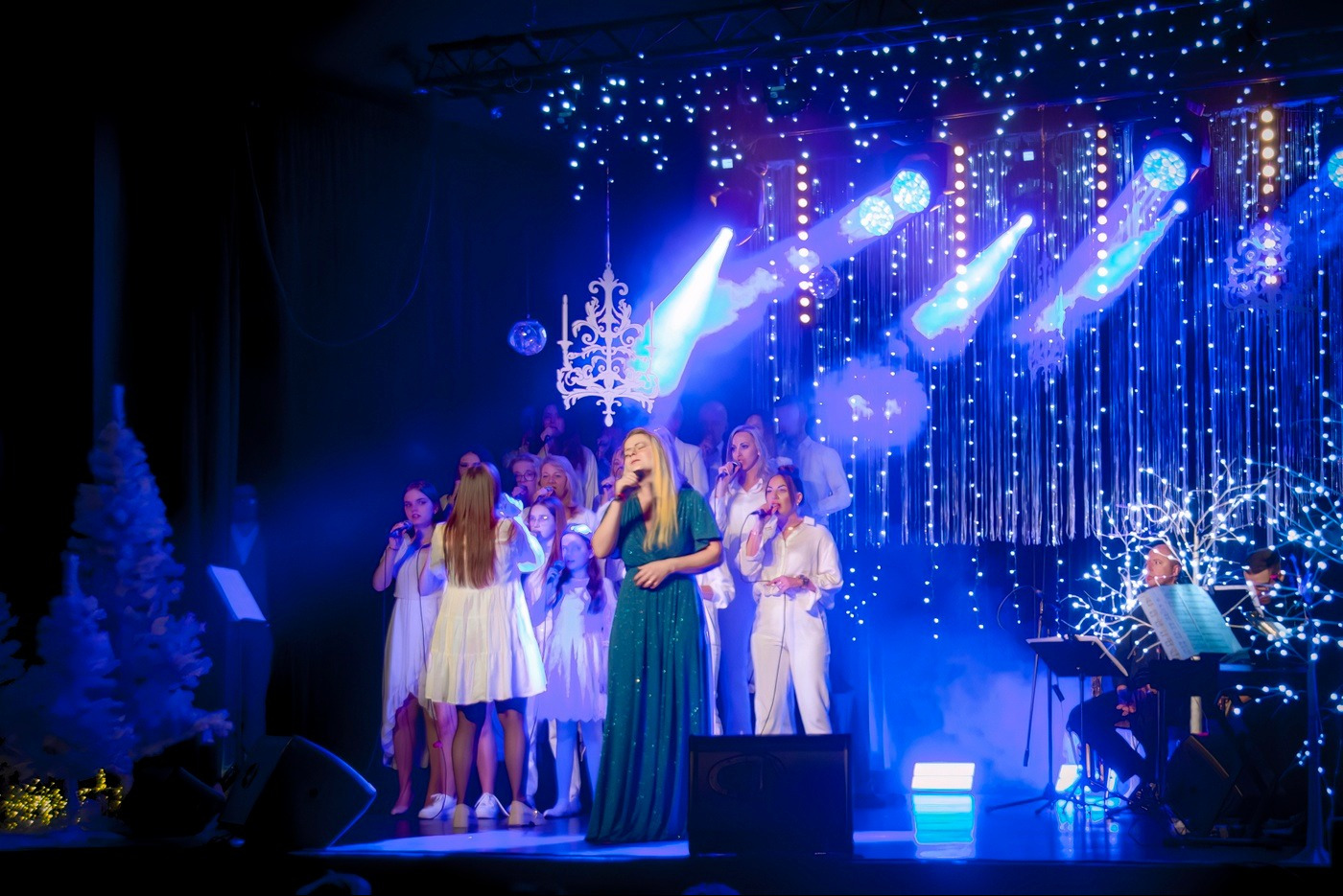 Chór na scenie podczas występu świątecznego, oświetlenie punktowe, dekoracje z lampek, wokalistka w szmaragdowej sukni na pierwszym planie.